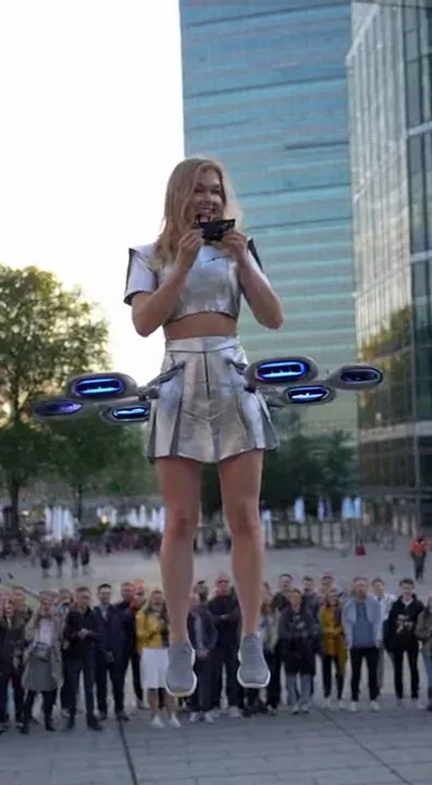 beautiful European woman in a short futuristic skirt and short futuristic top fly about 5 meters above ground before a curious crowd in a modern city square. Around her waist is a sleek personal flight device with small aerodynamic propellers glowing softly. She holds a compact remote controller in her hands, smiling confidently as she demonstrates how the device allows her to lift gently off the ground and hover gracefully in midair. She really fly!!! The dron like real with 6 propellers

The audience watches in awe as she explains the concept of individual flight technology. Sunlight reflects off nearby glass buildings, subtle wind effects stir her hair and skirt, and the camera circles around her dynamically. Cinematic science fiction style, ultra-detailed lighting, realistic physics, 4K resolution, immersive sound with crowd reactions and mechanical hums.” And at the end we can pigeon 