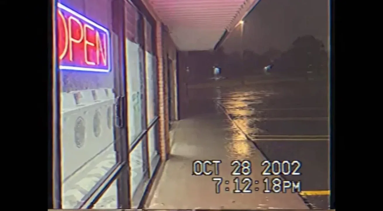 Real life Authentic raw VHS camcorder footage from the 2000s, recorded directly onto magnetic tape — not playback on a TV. The image has soft blur, muted colors, analog noise, faint static, and occasional horizontal tracking lines near the bottom. Slight handheld camera motion with natural jitter. Subtle chroma bleeding, color drift, and scanline flicker give it a genuine analog feel. Timestamp overlay with seconds in small white digital text is locked in the bottom-right corner, perfectly stable and unaffected by camera movement, static, or distortion. The timestamp never glitches, warps, or disappears — it remains consistently visible throughout. The audio has a low hiss, faint tape hum, and distant ambient noise captured from the camcorder’s built-in microphone. Looks exactly like genuine VHS footage, not a digital filter — 📼 “OCT 28 2002 7:12:18 PM – home video of a stormy night outside a laundromat, rain hitting pavement, the neon ‘OPEN’ sign flickers, camera pans toward the empty parking lot.” 🅿️